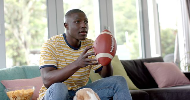 Young man is sitting on sofa, holding football while watching game, possibly on television. Image can be used for themes related to sports, home leisure activities, football fans, or promotional content for sporting events and at-home entertainment.