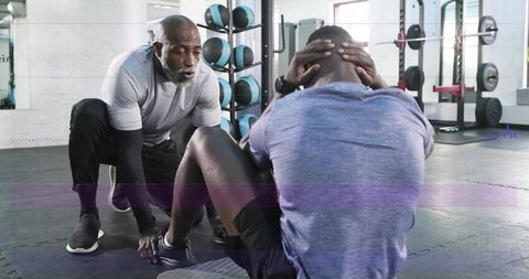 Trainer bracing client feet while client doing sit-ups on mat in modern gym with weights