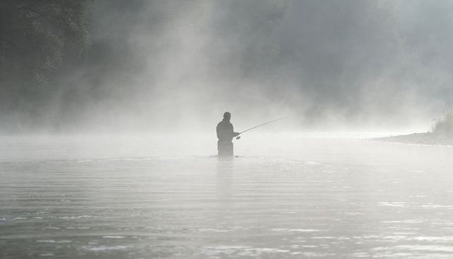 Solitary angler standing waist-deep in misty river casting rod at dawn for calm outdoors