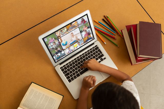 Child engaged in online virtual learning through a laptop. Various colored pencils and books nearby indicate active study and engagement. Suitable for illustrating concepts related to remote education, virtual classrooms, or the use of technology in modern education frameworks. Ideal for educational websites, online learning platforms, or articles about remote learning trends.