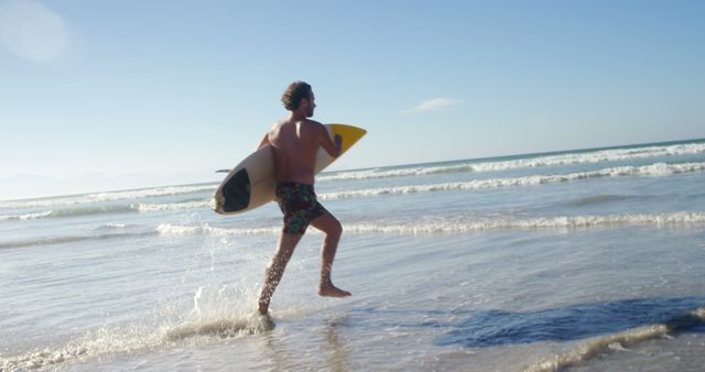 A young Caucasian man runs towards the ocean waves with a surfboard, ready to enjoy a surfing session. His enthusiasm for the sport is evident as he prepares to catch the waves under the clear blue sky.