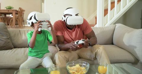 African American Father and Son Playing VR Headsets Together on Living Room Sofa