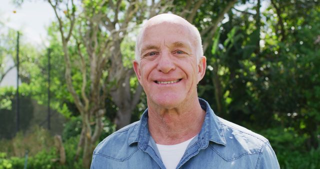 Senior man smiling in sunny backyard garden. Seeming happy and relaxed in casual attire. Perfect for use in materials related to retirement living, outdoor activities, gardening, or promoting a positive lifestyle for seniors.