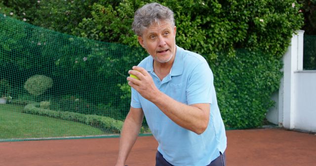 Senior man practicing tennis on outdoor court, holding tennis ball and preparing for serve. He appears active and engaged in a healthy lifestyle. Suitable for topics about fitness, sports, aging, health, and outdoor activities.