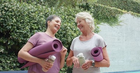 Two women laughing after outdoor yoga holding purple mats and water bottles