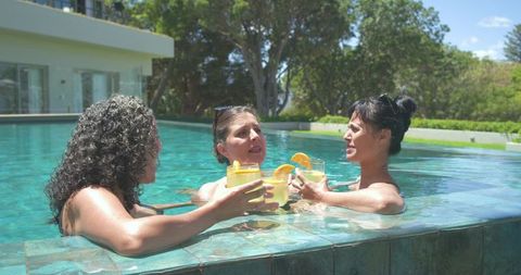 Three women toasting at pool ledge with citrus cocktails and sunglasses, backyard leisure