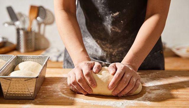 Hands kneading dough on wooden countertop wearing flour-dusted apron with loaf pan