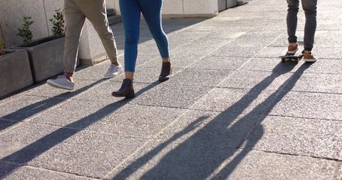 Friends Walking and Skateboarding on Sunlit Urban Sidewalk Casting Long Shadows