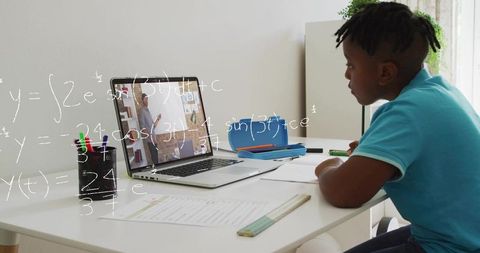 Boy studying online math lesson at home desk watching teacher on laptop with worksheet
