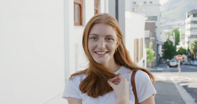 Young redhead woman smiling while standing on city street. Casual clothing suggests relaxation and friendliness. Ideal for use in blogs or advertisements promoting urban lifestyle, outdoor activities, or youth-centric products.