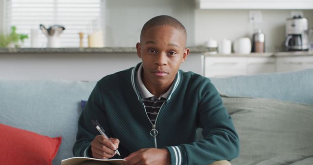 Focuses on African American teenage boy seated on sofa, engaging in study or homework with notebook and pen. Bright, modern living room and kitchen background suggests a home environment. Ideal for educational themes, academic success, remote learning resources, youth studies, tutoring services.