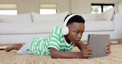 Boy lying on rug using tablet and wearing headphones at home focusing on screen