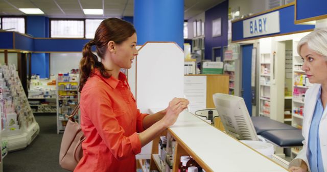 A woman stands at the pharmacy counter asking the pharmacist for health advice. She is wearing a red blouse and carrying a bag. The pharmacy is well-lit and organized, displaying shelves with various products. This visual can be useful for illustrating healthcare services, customer consultations, medical advice scenarios, pharmacy interior layouts, and patient-pharmacist interactions.