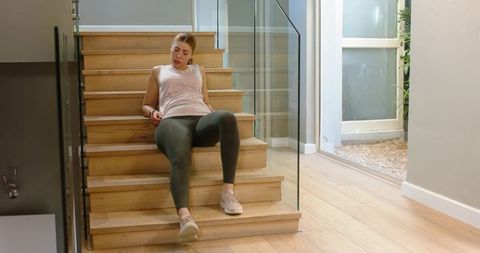 Woman resting on wooden stairs after workout with floating fitness metrics in modern home