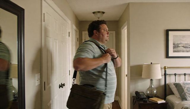 Man adjusting shoulder bag strap in bedroom hallway wearing striped polo and khakis