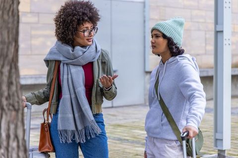 Two diverse women chatting with rolling luggage in city plaza wearing scarves and beanies
