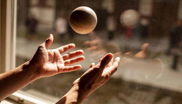 Cradling hands tossing wooden sphere near sunlit window with warm bokeh and reflection
