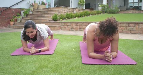 Diverse women holding forearm plank on backyard lawn with purple mats and fitness watches