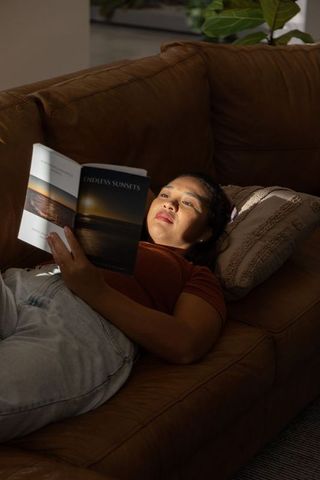 Asian woman reclining on leather sofa reading 'endless sunsets' book in warm sunlight