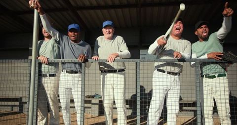 Baseball teammates cheering and celebrating in dugout leaning on fence waving bat and glove