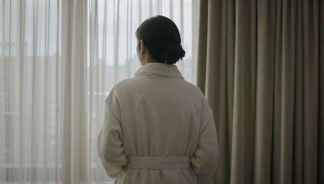 Serene asian woman standing at window wearing belted robe in soft daylight