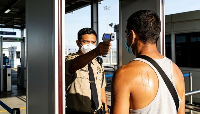 Security guard checking forehead temperature of masked traveler at airport checkpoint