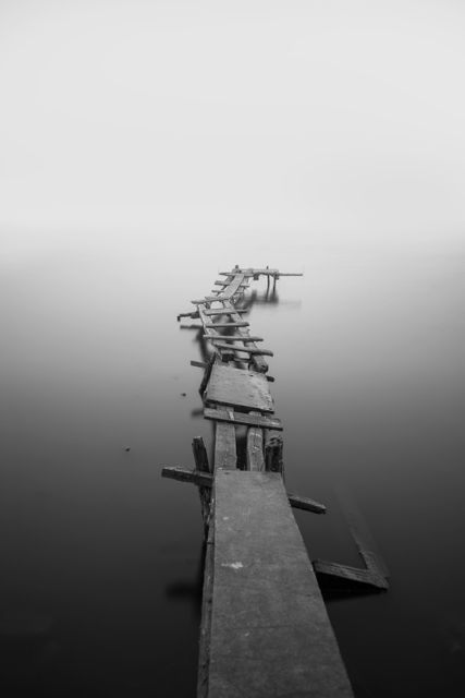 An old, broken wooden bridge extending over calm, misty water. Perfect for concepts of solitude, abandonment, tranquility, or mystery. Ideal for use in nature-themed designs, backgrounds, and inspirational contexts.