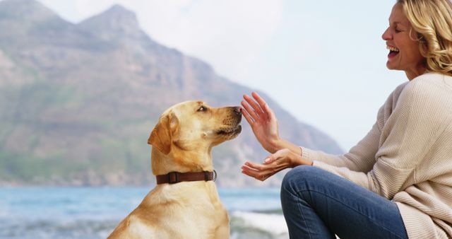 A middle-aged Caucasian woman enjoys a playful moment with her Labrador Retriever on a beach, with copy space. Her laughter and the attentive stance of the dog create a warm, engaging atmosphere.