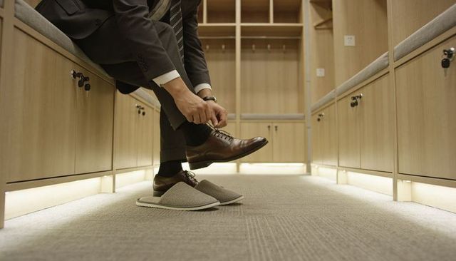 Man tying leather shoe on locker room bench with slippers, woodgrain cabinetry and soft light