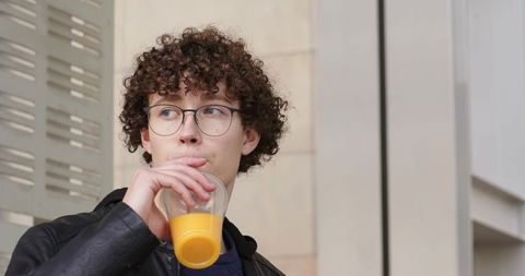 Young man sipping orange juice on city sidewalk wearing leather jacket and glasses