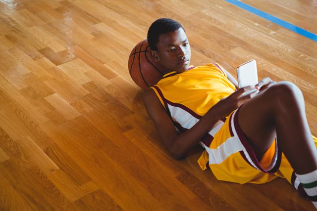 Teenage basketball player in yellow uniform lying on hardwood floor, using mobile phone. Ideal for themes related to youth, sports, technology, leisure, and athletic lifestyle. Can be used in articles, advertisements, or social media posts about teenage life, sports activities, or mobile technology.