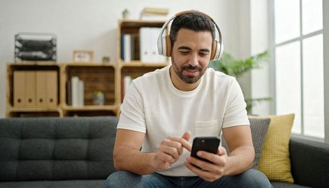 Man wearing headphones using smartphone on sofa in bright minimal living room with natural light