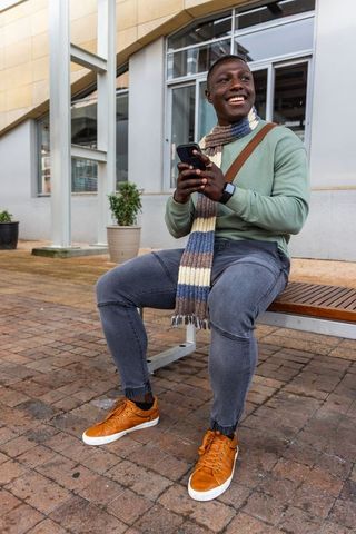 Smiling african american man sitting on bench holding phone in modern urban plaza wearing scarf