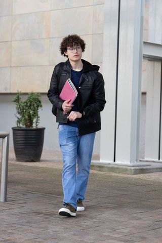 Young male student walking on campus holding pink folder, wearing black jacket