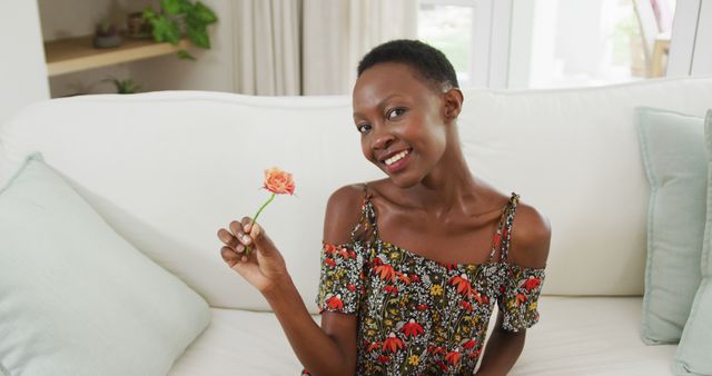 African american woman sitting on sofa smelling flower and smiling to camera. staying at home in isolation during quarantine lockdown.