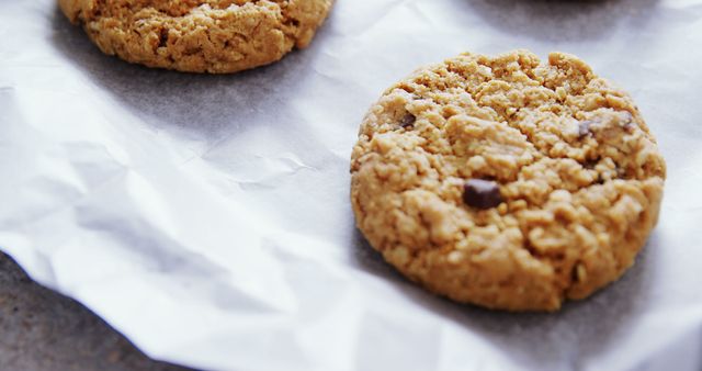 Freshly baked chocolate chip cookies rest on parchment paper, with copy space. Their golden-brown texture and scattered chocolate chunks invite a moment of indulgence.