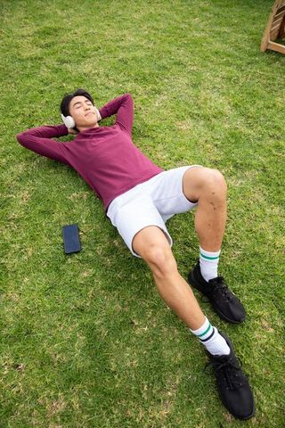 Young asian man relaxing on green lawn listening to music with wireless headphones