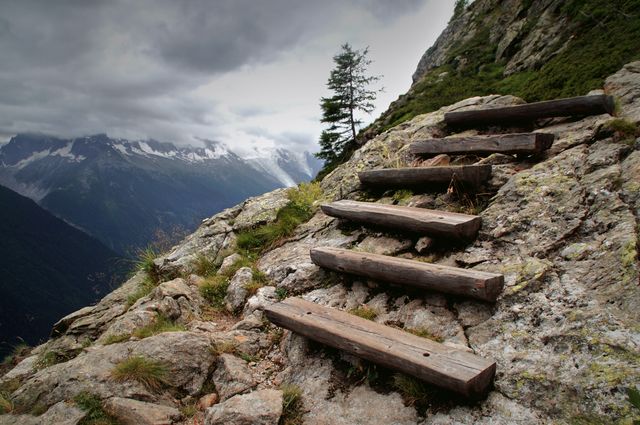 Wooden steps installed on a rocky mountain path leading to a panoramic view of distant mountain ranges. Perfect for use in travel blogs, adventure magazines, promotional material for hiking tours, and nature inspiration posters.