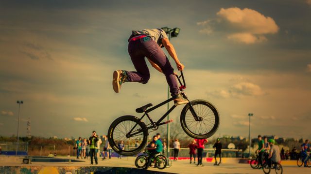 Young BMX rider executing a mid-air stunt at an outdoor skatepotencing park, with people observing in the background. Perfect for promoting extreme sports, youth activities, sportswear, urban lifestyle campaigns, and fitness inspiration.