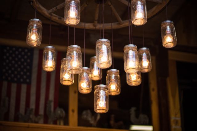 Rustic chandelier made of mason jars hangs in a cozy interior, casting warm light. Background includes an American flag, adding a touch of patriotic decor. Suitable for illustrating themes of country-style home decor, vintage ambiance, American heritage, and cozy interiors. Ideal for use in interior design blogs, patriotic advertisements, or farmhouse-themed promotions.