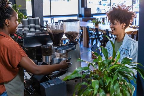 Barista preparing espresso while customer paying with phone at sunlit cafe counter