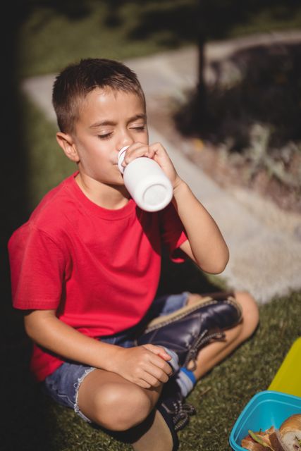 Young boy in red shirt and denim shorts drinking juice from a bottle while sitting on grass in schoolyard. Ideal for use in educational materials, health and nutrition campaigns, and advertisements promoting healthy lifestyles for children.