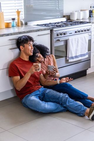 Diverse couple enjoying morning coffee on kitchen floor, leaning against cabinets