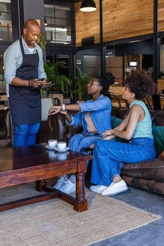 Cafe server taking order from two women on leather sofa using tablet in casual lounge