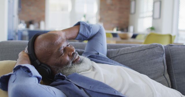 A mature man is relaxing on a comfortable couch indoors, enjoying music through his headphones. He appears to be happy and content, holding his hands behind his head and smiling slightly. This image can be used in advertisements for wellbeing, retirement, leisure products, senior living, health and relaxation concepts.