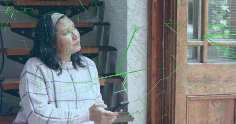 Mature woman holding smartphone gazing toward window light with green digital overlay