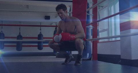 Squatting boxer resting in ring corner wearing red gloves and black shorts after intense round