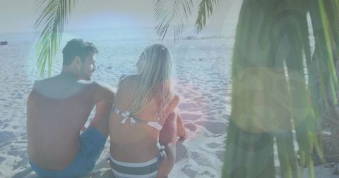 Couple sitting on sandy beach facing ocean under palm fronds with sunlit lens flare