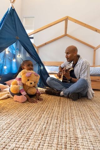 Father playing guitar with daughter in cozy bedroom teepee hugging teddy bear on woven rug