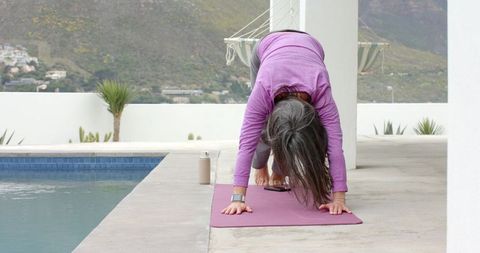 Silver-haired woman practicing yoga by pool on terrace, stretching on magenta mat